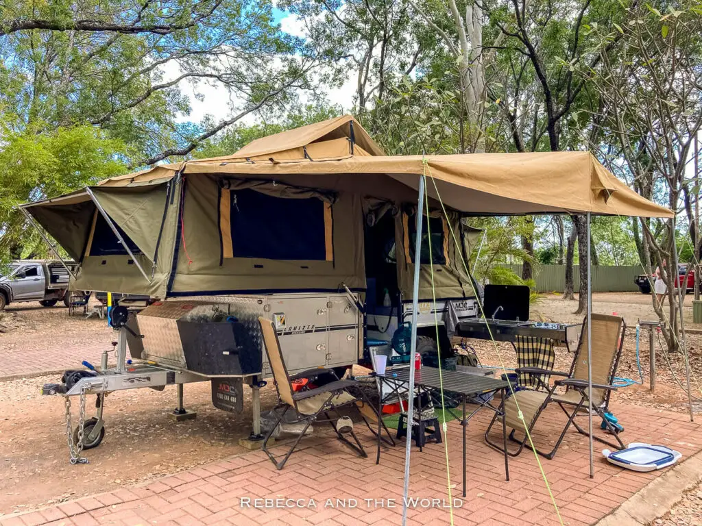 A camping trailer with an attached canvas tent is set up outdoors. The tent is extended over a picnic table with chairs, cooking equipment, and various camping supplies. Trees and another vehicle are visible in the background.