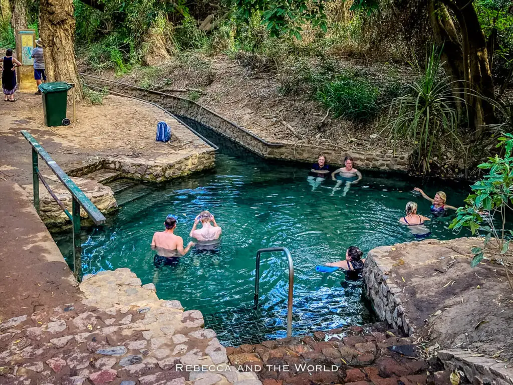 People are relaxing in a natural swimming hole - the Katherine Hot Springs - surrounded by a stone ledge with a metal handrail leading into the water. Lush greenery and trees envelop the area, providing a serene backdrop.