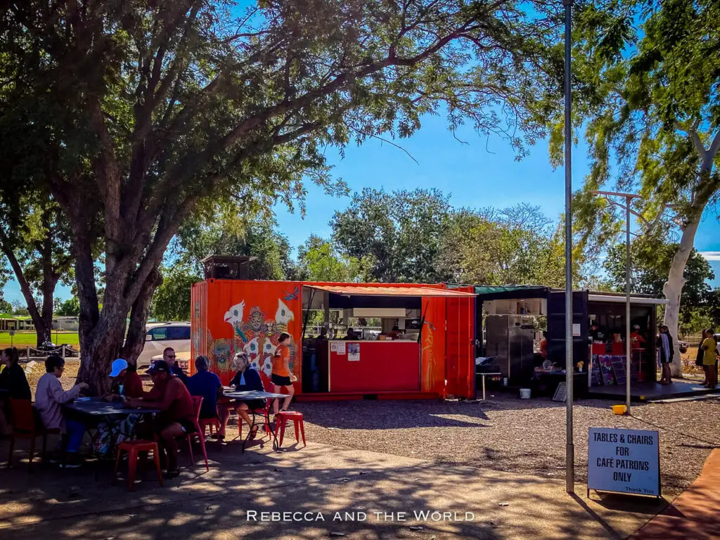 Patrons are seated at red tables outside the "Pop Rocket Cafe" made from a shipping container. Trees provide shade overhead, and a sign indicates seating is for café customers only. The setting is casual and welcoming.