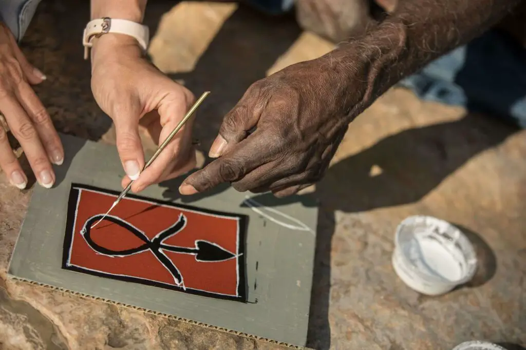 A close-up of two people's hands working on a painting. The person is using a thin, pointed tool to apply white paint onto a dark, rectangular surface with a red background, creating an abstract design. A small, open container of white paint is visible to the side. The hands appear weathered and experienced, suggesting this may be the work of an artisan. Top Didj is a great cultural experience in Katherine, Northern Territory.