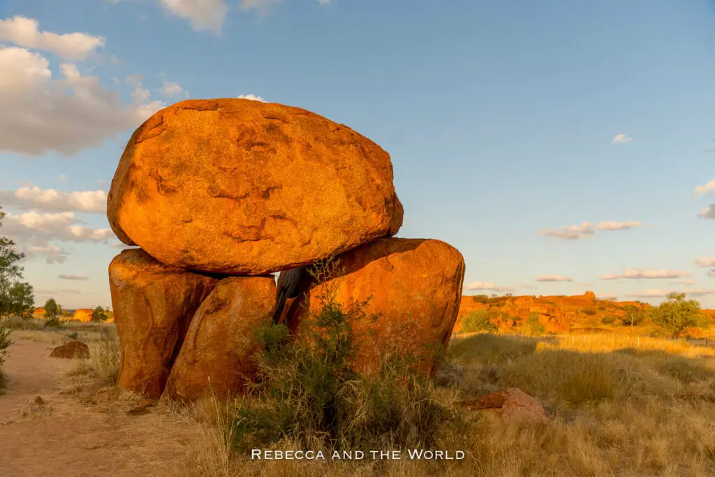 A large, balanced rock formation stands prominently in the foreground against a clear blue sky with soft clouds. The warm golden light of the setting or rising sun illuminates the red-orange surface of the rocks, highlighting their textures. In the background, more rock formations dot the landscape, interspersed with sparse vegetation and dry grasses. This is Karlu Karlu, or Devils Marbles, one of the must-see Stuart Highway attractions.