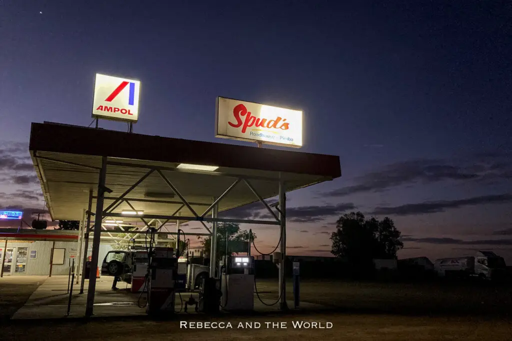 Night-time scene at a roadside service station on the Stuart Highway with illuminated signs for Ampol and Spud's Roadhouse, and a dark sky in the background.