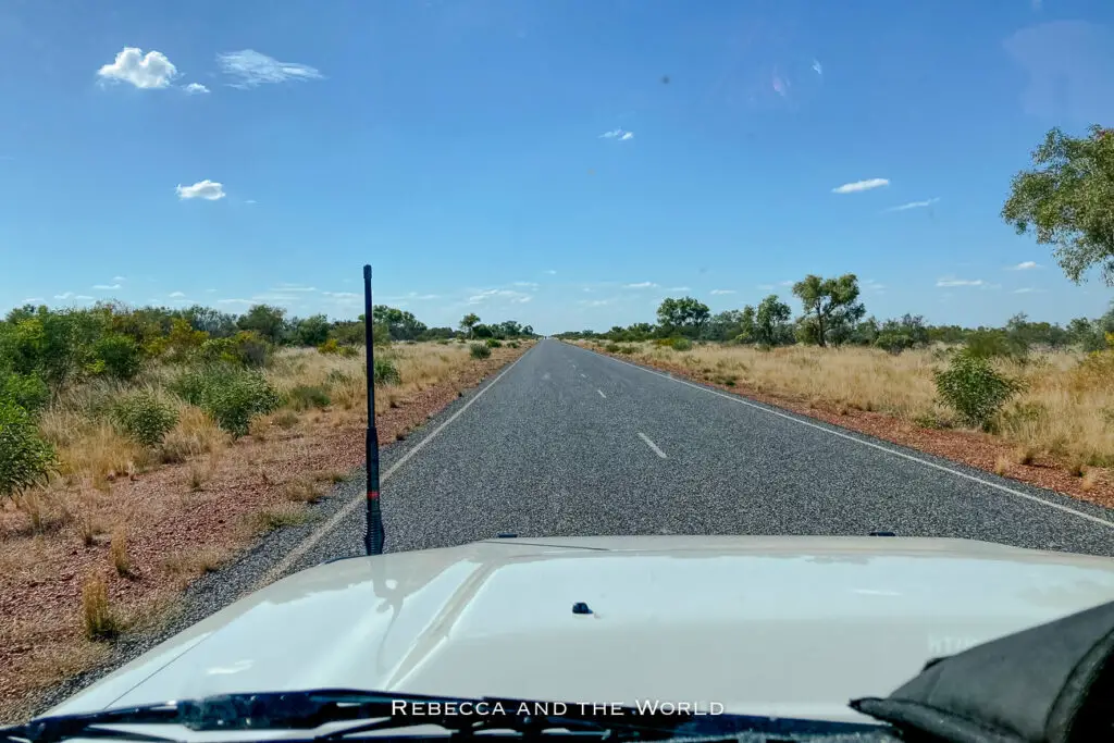 Driver's perspective of a long straight road - the Stuart Highway - stretching through sparse Australian outback with bushland on either side and a clear blue sky overhead.