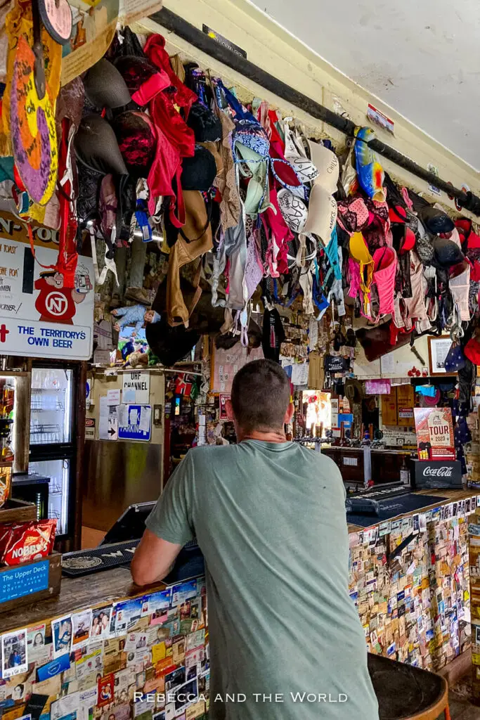 Interior of a rustic Australian pub with a collection of bras hanging from the ceiling and various notes pinned to the walls, with a man - the author's husband - ordering at the bar. This is the famous Daly Waters Pub, a must-visit along the Stuart Highway on am Adelaide to Darwin road trip.