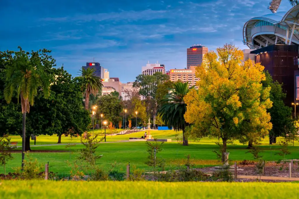 View of Adelaide's skyline at twilight with lush greenery in the foreground, showcasing a blend of modern buildings and abundant parkland. Adelaide is the perfect starting point for an Adelaide to Darwin road trip!