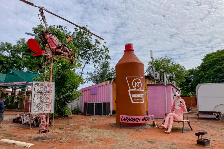 Quirky roadside attraction featuring a large homemade bottle sculpture and a pink panther figure sitting on a bench, with a mock helicopter overhead. Stop at the Larrimah Hotel on your Adelaide to Darwin road trip itinerary.