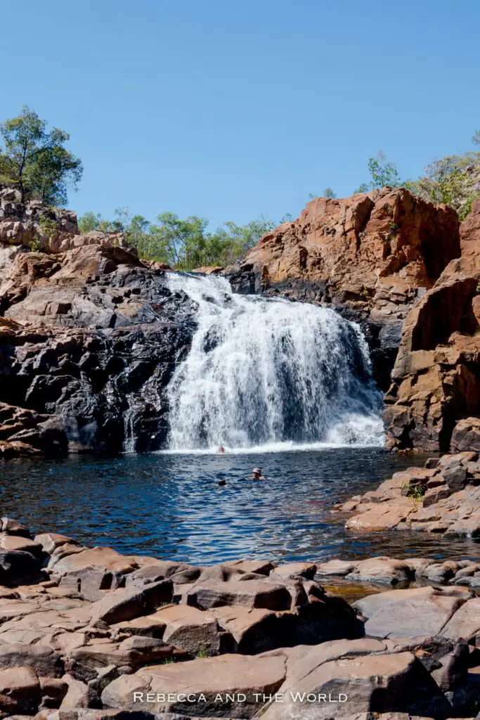 A wide-angle view of a waterfall with water flowing over a stepped rock formation into a clear blue pool. Reddish-brown rocks and sparse green vegetation surround the area. Swimmers are visible in the water, enjoying the refreshing cascade. This is Leliyn Falls in beautiful Nitmiluk National Park in the Northern Territory.