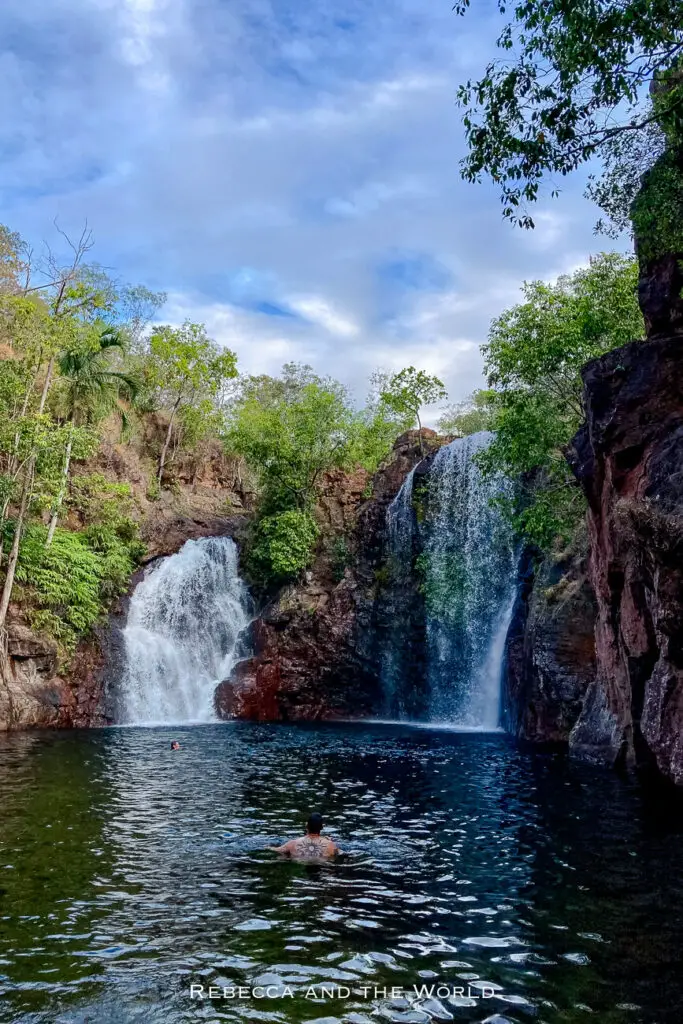 A serene natural pool at the base of a double waterfall, flanked by red rocky cliffs and green vegetation. The sky is partly cloudy with blue patches. People can be seen swimming in the calm water, enjoying the natural surroundings. This is Florence Falls in Litchfield National Park in the Northern Territory.