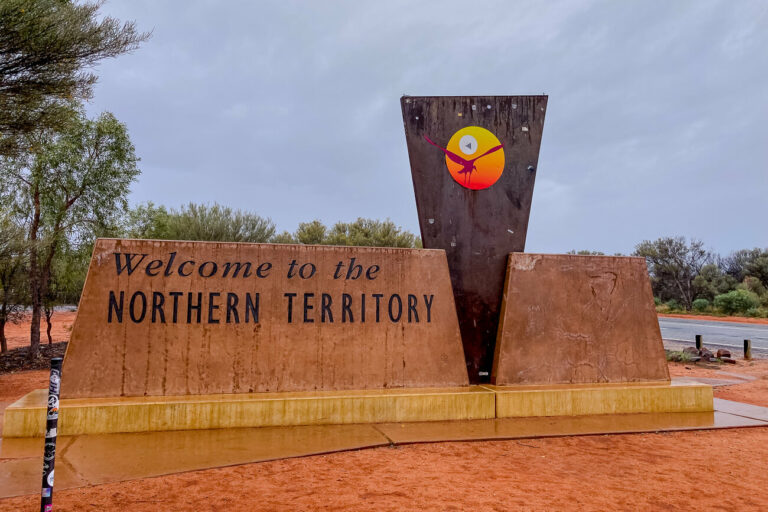 A roadside welcome sign reading "Welcome to the Northern Territory" in large lettering on a rust-coloured backdrop. The sky is overcast, and the ground is the iconic red dirt of the Australian Outback.