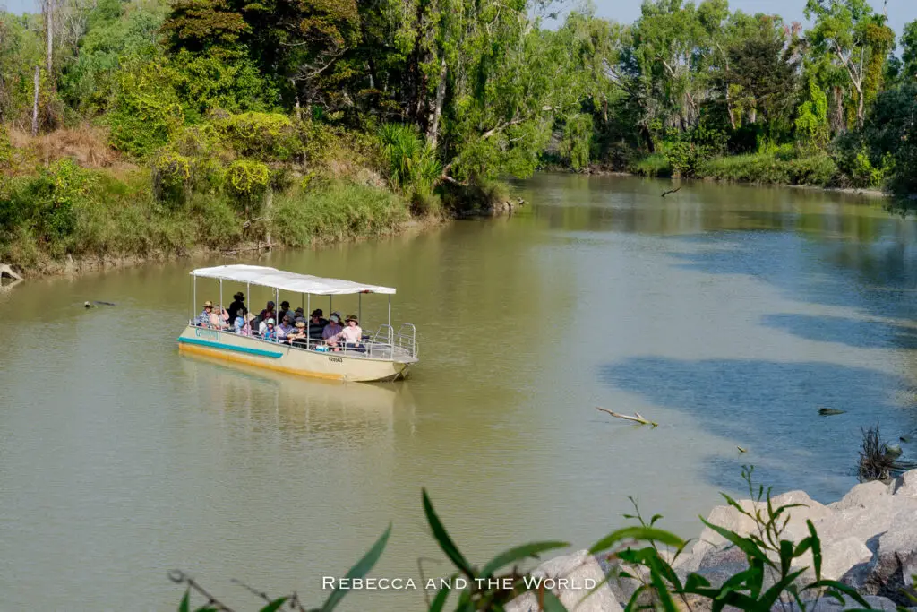 A riverboat with a canopy top carrying passengers on a calm river, flanked by lush greenery on both banks. The Guluyambi Cultural Cruise is a great way to learn about the Indigenous Traditional Owners of Kakadu and the importance of the national park.