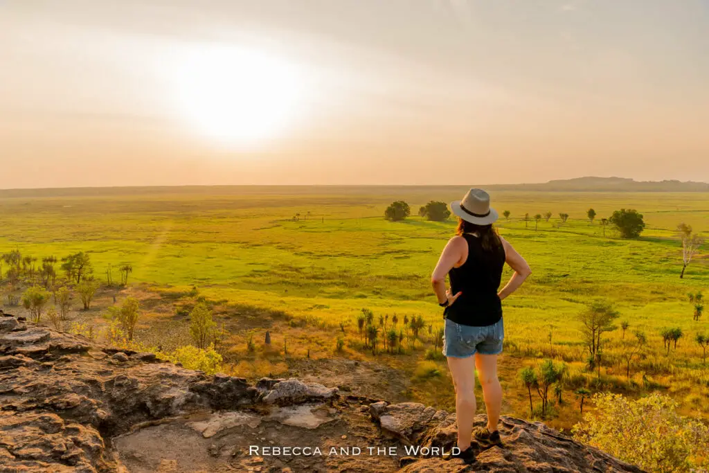 A woman - the author of this article - standing on the rocky ledge of Ubirr in Kakadu National Park overlooking a vast savannah under a hazy sky, the sun low on the horizon.