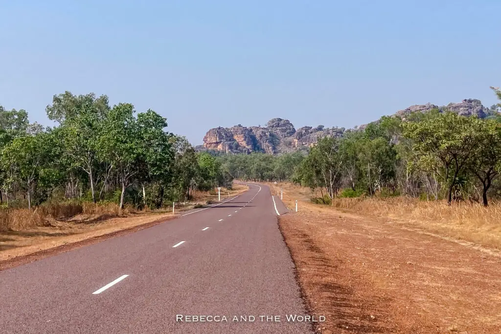 A straight road leading towards distant rock formations, bordered by tropical savannah and a clear blue sky. The road leads to Burrungkuy, one of the most significant sites in Kakadu National Park.