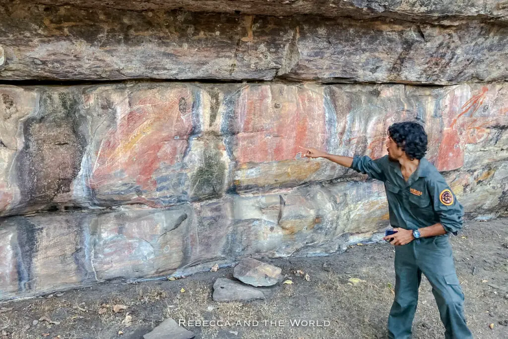 An indigenous guide pointing to ancient rock art on a sheltered stone wall at Ubirr in Kakadu National Park, displaying various traditional paintings.