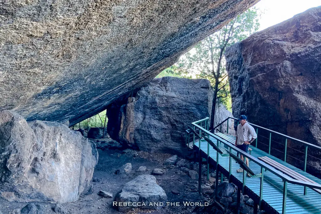 A metallic walkway leading through a rocky terrain with large boulders and trees, partially shaded by the overhanging rock. This is Burrungkuy in Kakadu National Park, one of the most significant sites in the park.
