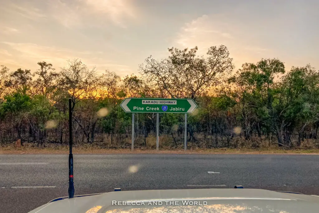 Road sign indicating directions to Pine Creek and Jabiru on Kakadu Highway at dusk with the silhouette of a car antenna in the foreground and a sunset sky in the background.