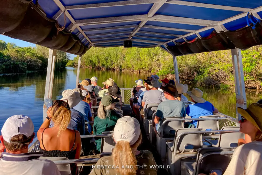 View from the rear of a covered riverboat filled with tourists wearing hats, cruising on a calm river bordered by dense greenery in Kakadu National Park. The Yellow Water Cruise is one of the best things to do in Kakadu National Park.