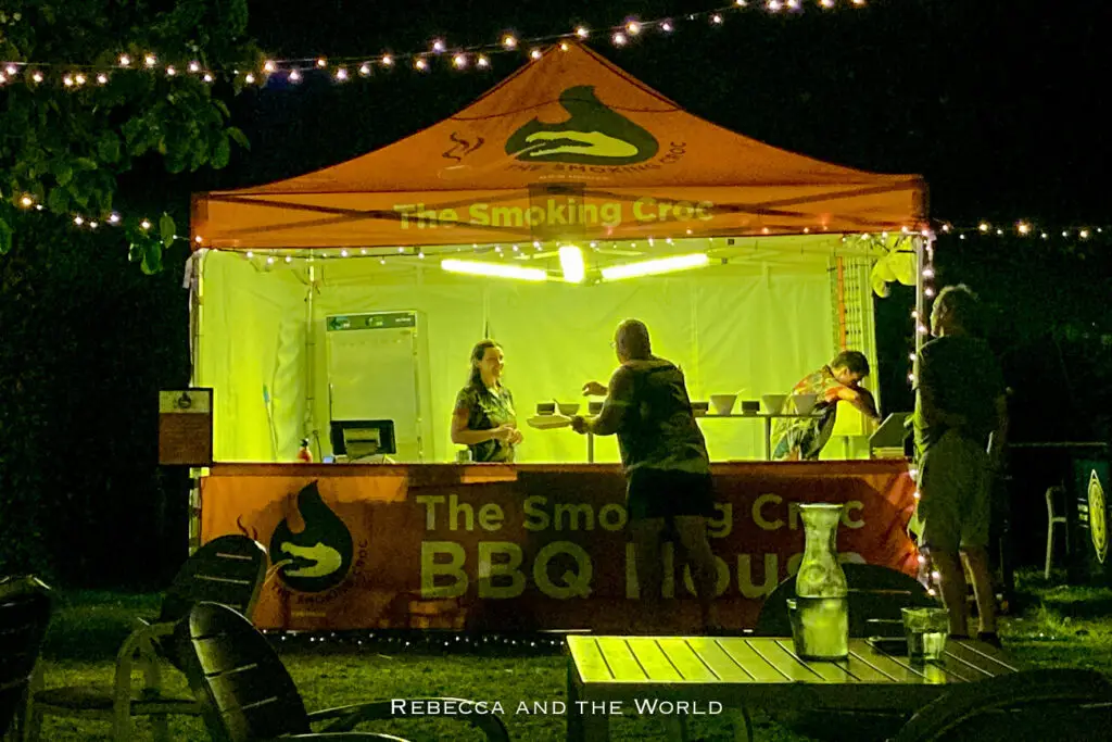 A night view of a food stall named 'The Smoking Croc BBQ House' under a tent lit by string lights, with vendors serving customers at a counter. Cooinda Lodge is really the only place inside Kakadu National Park where you can get food.