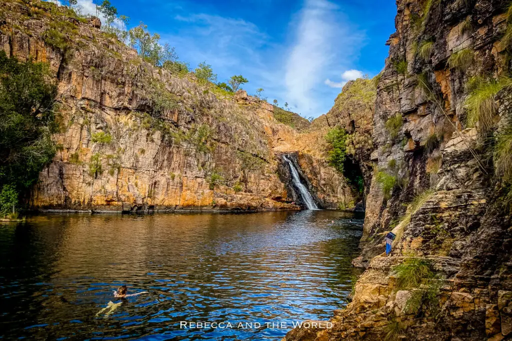 The Beautiful Maguk Falls in Kakadu National Park cascading into a tranquil natural pool surrounded by rugged cliff walls with vegetation, while visitors enjoy a swim.