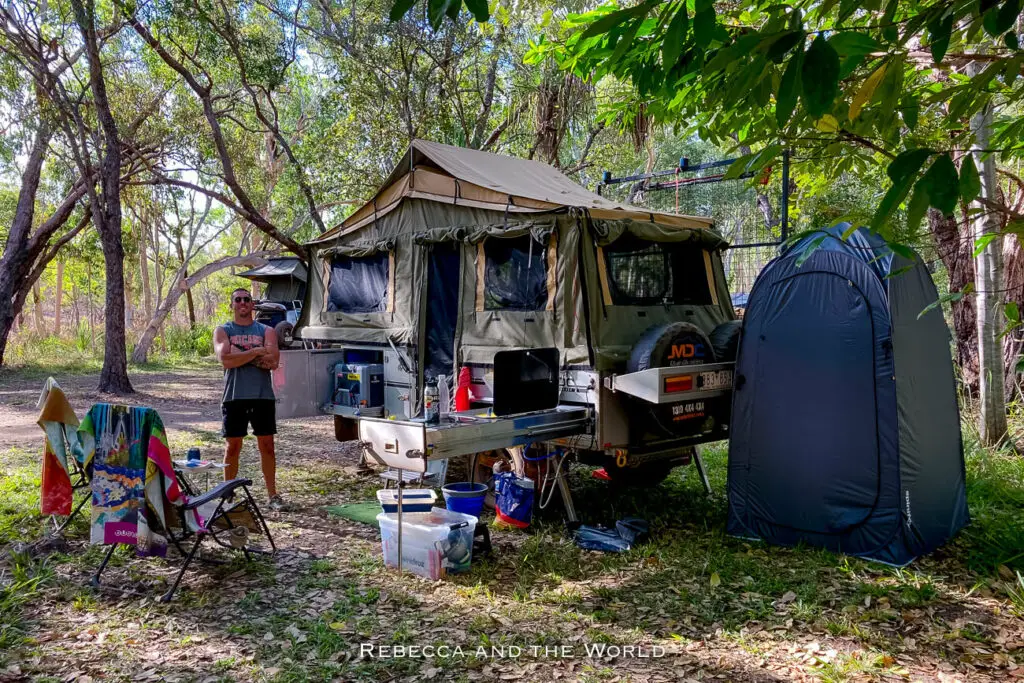 A man standing beside a camping setup in the woods with a tent mounted on a 4WD vehicle and camping chairs and equipment arranged around. This is Maguk campground in Kakadu National Park.