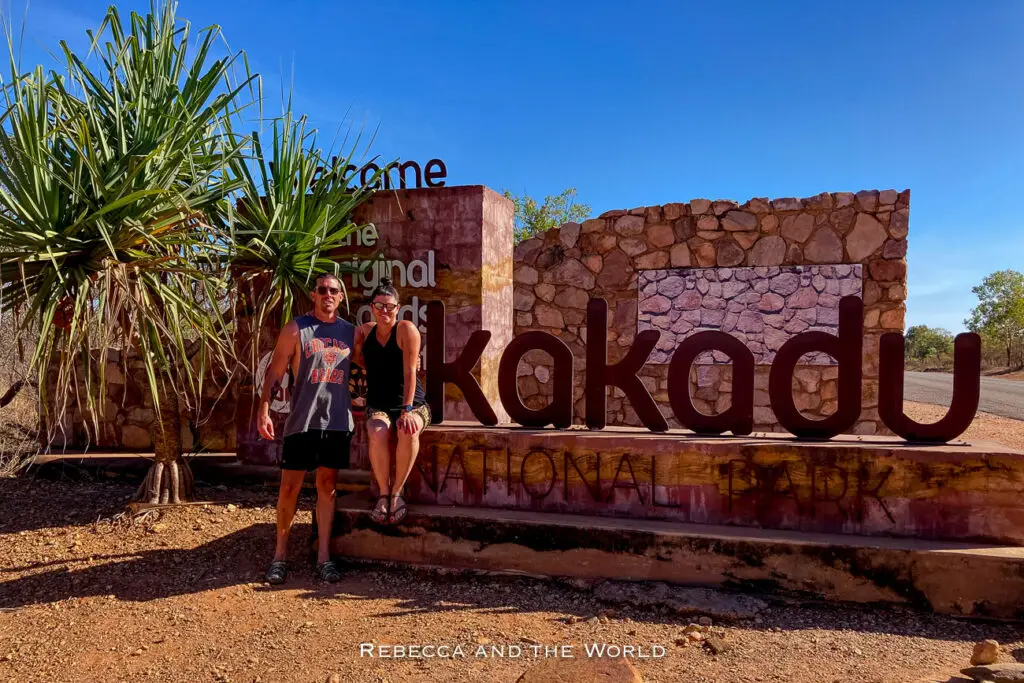 Two people - the author and her husband - smiling and posing in front of the large, iconic sign at the entrance of Kakadu National Park, with the text "Kakadu" prominently displayed in large, three-dimensional letters.
