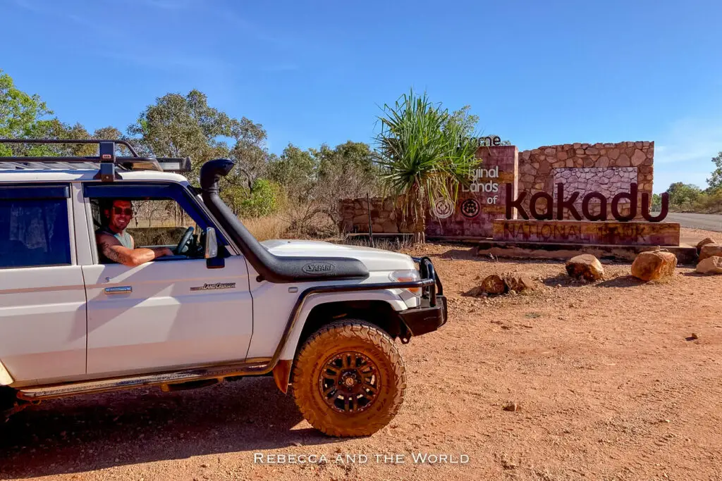A four-wheel drive vehicle parked at the entrance of Kakadu National Park, with a sign reading "Welcome to the Aboriginal Lands of Kakadu National Park" in the background. A man - the author's husband - smiles at the camera from the driver's seat.