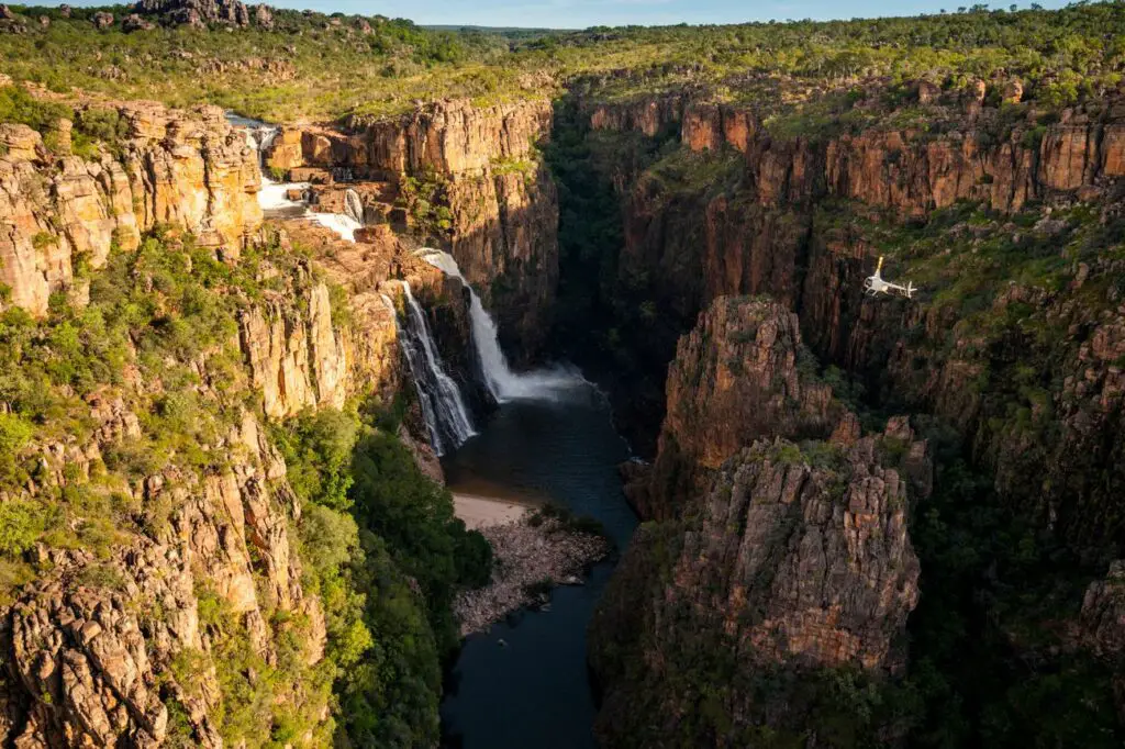 The powerful Twin Falls cascade down a sheer cliff face in Kakadu National Park, creating a misty spray that catches the sunlight, forming a delicate rainbow across the gorge's depth, symbolising the park's pristine beauty and serene atmosphere. A helicopter flies near the falls.