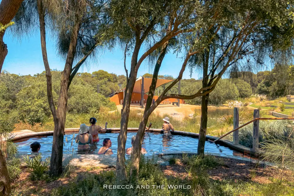 Group of people enjoying a hot spring surrounded by native trees and shrubbery, with a rustic building in the background. The Peninsula Hot Springs is one of the best places to visit in Victoria.