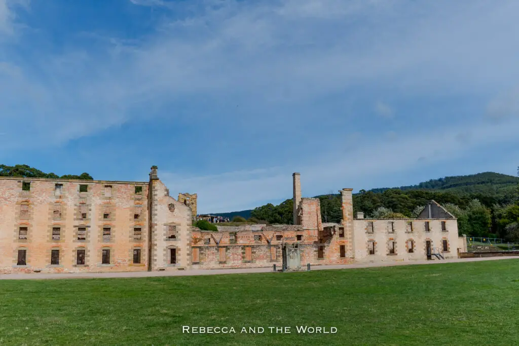 Historic ruins of a large brick-built penitentiary with multiple chimneys, grassy foreground, and a hill in the distance. One of the best things to do in Tasmania is learn about our colonial history at Port Arthur.