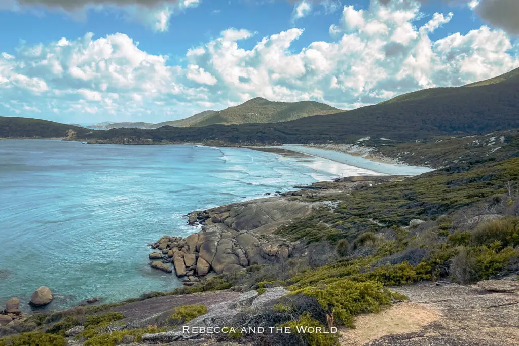 Scenic view of a pristine bay with turquoise waters, surrounded by rolling hills and a rugged coastline, under a sky dotted with fluffy clouds. This is Wilsons Promontory, one of the best places to visit in Victoria, Australia.