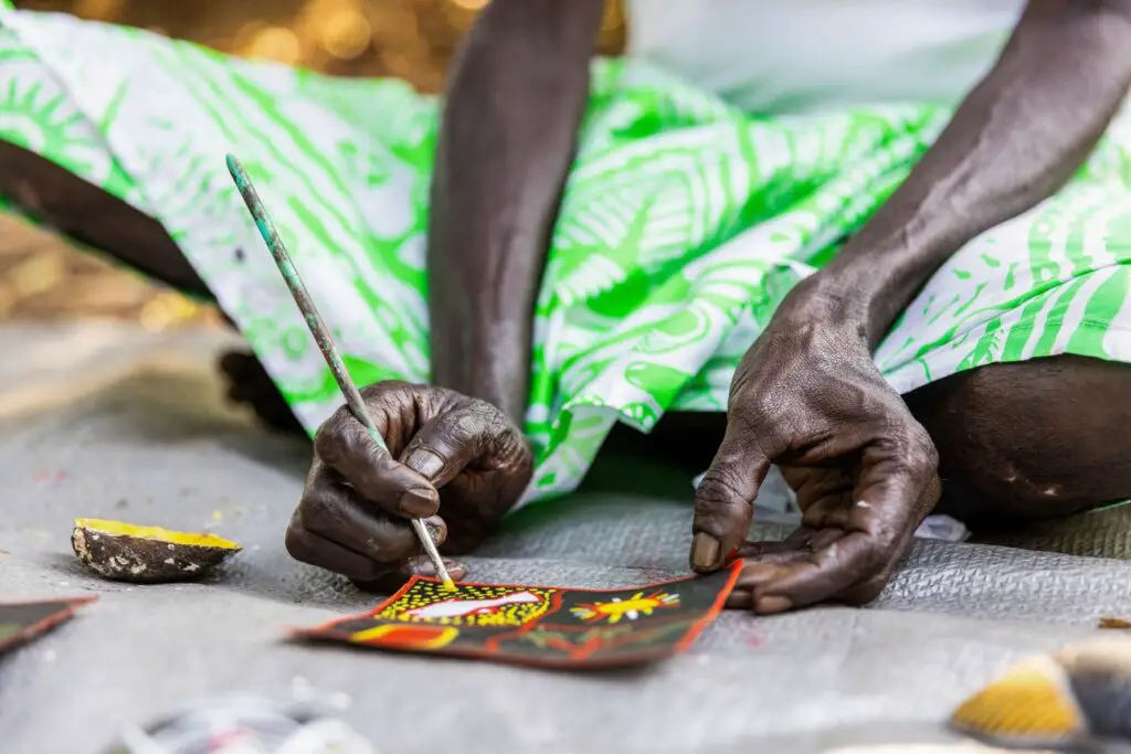 The focus is on the hands of a person painting intricate designs on a small rectangular canvas. The hands are dark and weathered, indicating a skilled artisan at work. The background is a blurred pattern of green and white fabric, which creates a contrast that highlights the detailed artwork and the concentration involved in the craft.