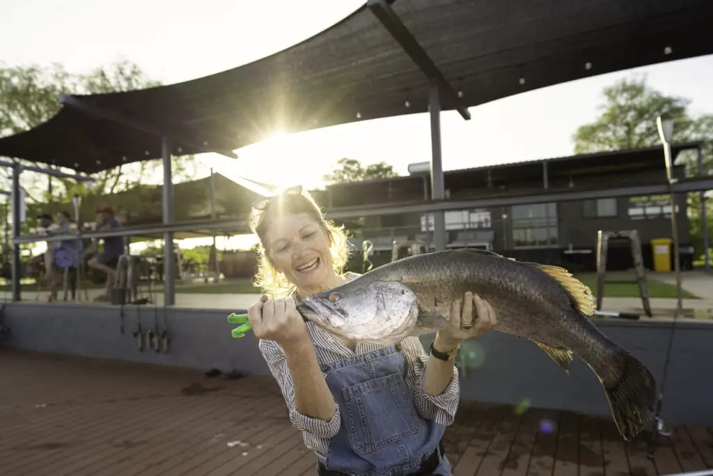 A woman in a denim overall and striped shirt smiles broadly while holding a large fish in both hands. She is outdoors, and the sun is setting behind her, casting a warm glow and causing lens flare. Behind her, there are people sitting at outdoor tables under a large shade structure. Going fishing is a must-do when you visit the Northern Territory!