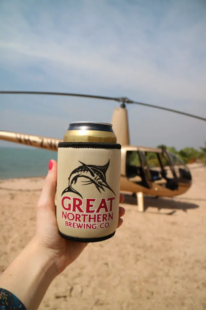 A close-up of a hand holding a can cooler with the logo "Great Northern Brewing Co." printed on it, featuring a marlin fish. In the background, there's a sandy beach and a helicopter, partially visible with its door open, suggesting a scenic tour or a remote location. One of the most luxury things to do in the Northern Territory is a helicopter pub crawl to remote pubs in the NT!