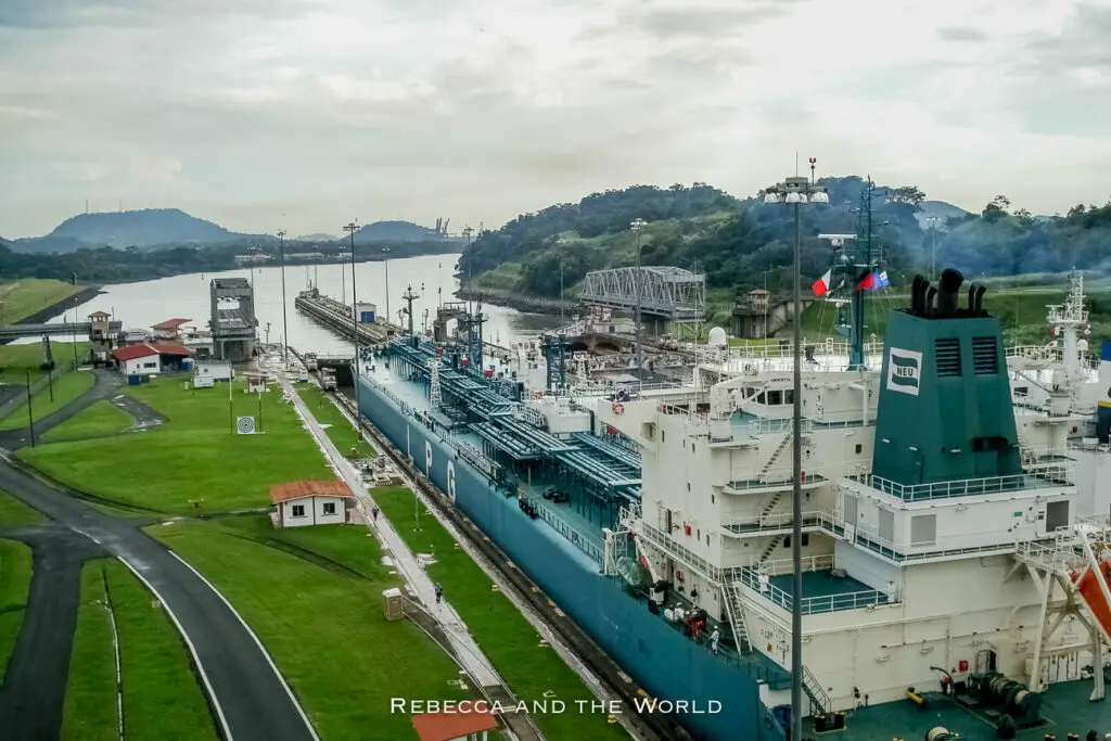 Overhead view of a large blue cargo ship passing through the Panama Canal. The ship is labeled with 'LPG' and the landscape features lush green hills in the background.
