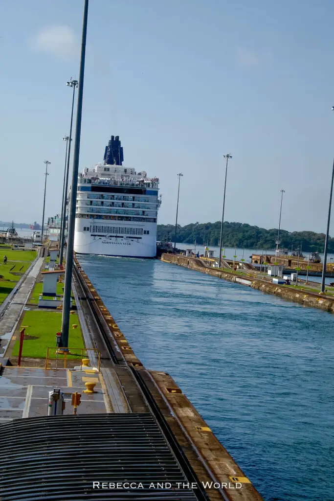 A large white cruise ship, the Norwegian Star, navigating through the narrow Panama Canal, with water on both sides and a clear blue sky overhead.