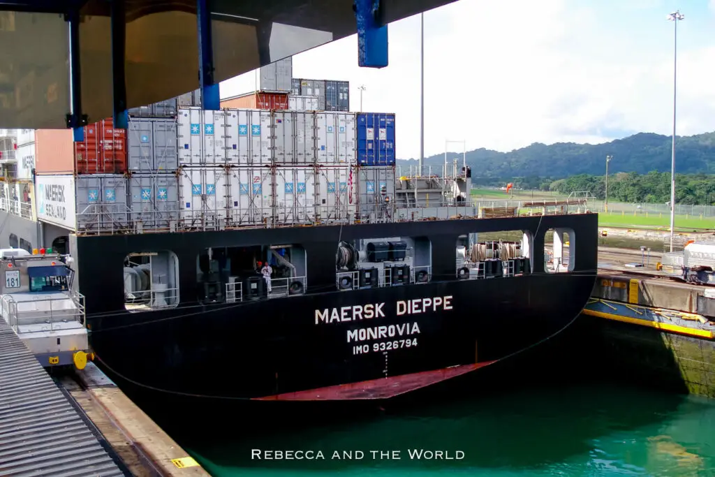 A large black cargo ship, the Maersk Dieppe, in the Panama Canal, filled with stacked containers of various colours, docked next to a canal lock.