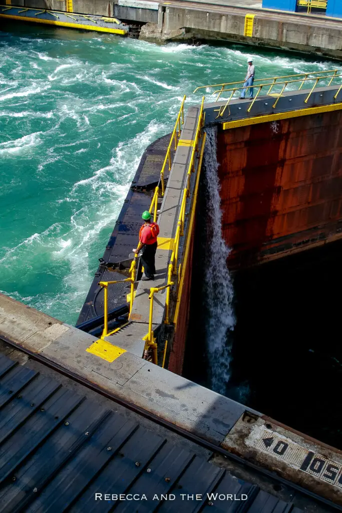 A canal worker in a red shirt and helmet stands on a narrow walkway next to the steel gate of the Panama Canal lock as water vigorously churns below.