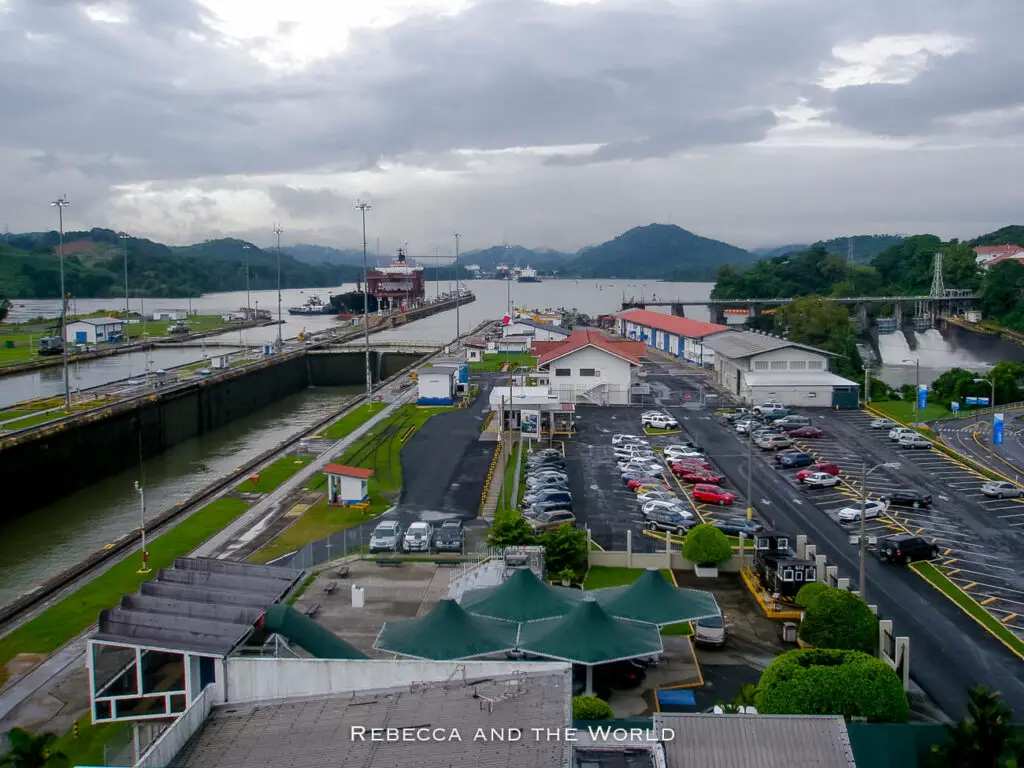 Aerial view of the Panama Canal lock system with a red cargo ship passing through. Surrounding the area are parking lots, buildings, and greenery under a cloudy sky.