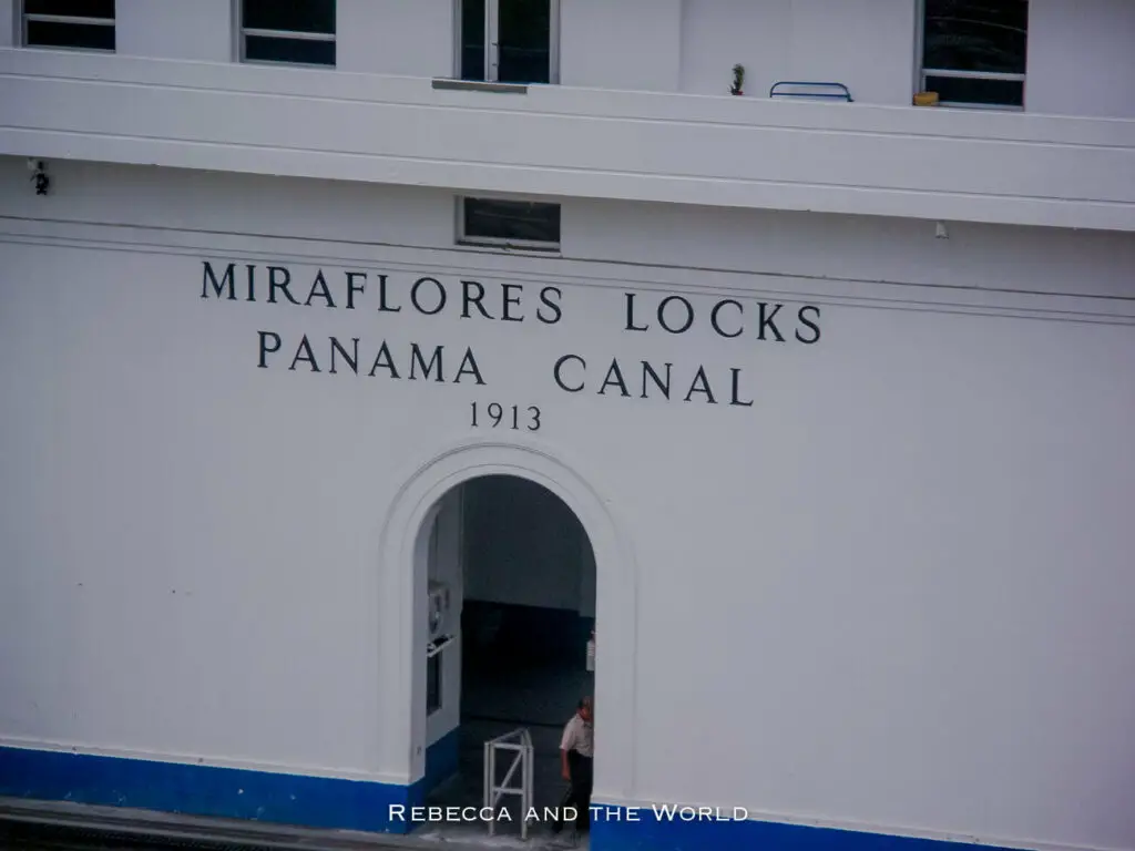 Close-up of a white wall with 'MIRAFLORES LOCKS PANAMA CANAL 1913' in bold black lettering, with an arched doorway showing interior details.