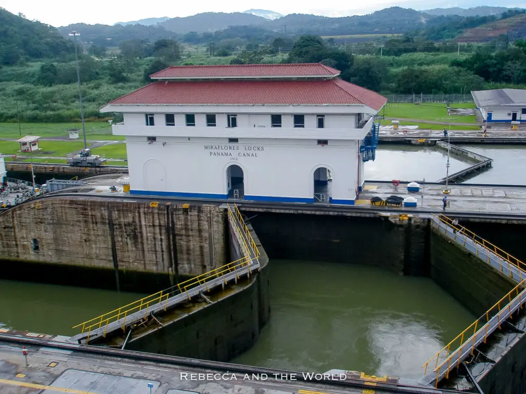 Elevated view of the Miraflores Locks at the Panama Canal, with a lock chamber filled with water, flanked by green lawns and administrative buildings against a hilly backdrop.