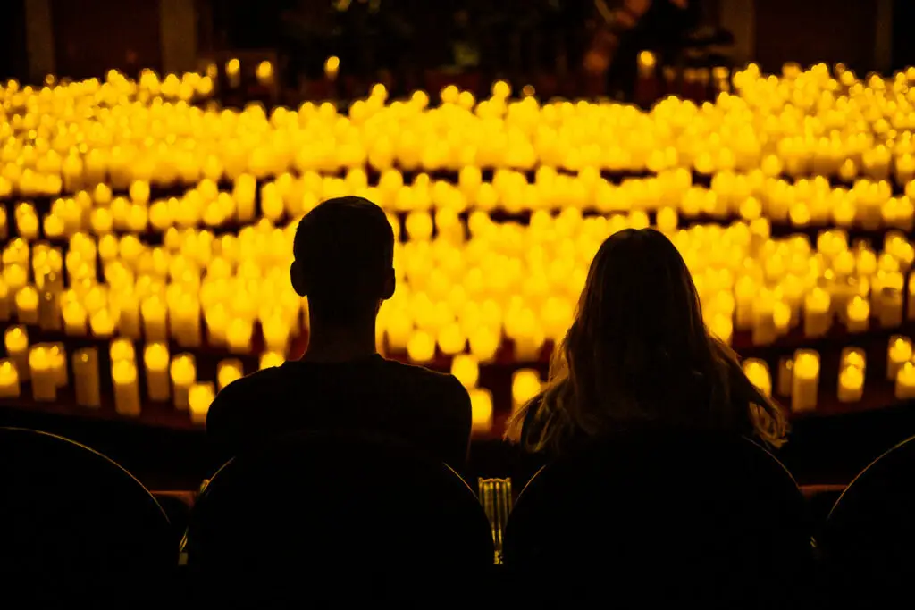 This image shows two individuals sitting in the foreground, viewed from behind, against a backdrop of numerous lit candles. The candles emit a warm, golden glow and are densely arranged, covering the entire background. The two individuals are in silhouette, which suggests they are in a dimly lit space, focused on the candles. Candlelight Concerts are one of the more unique things to do in Buenos Aires.