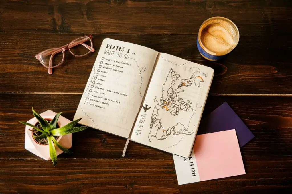 This image depicts a travel planning journal open on a wooden surface. On the left page, there's a checklist titled "PLACES I WANT TO GO" with various travel goals like "Trekking in South America," "Safari in Africa," and "Road trip in Scotland," some of which are checked off. The right page shows a world map with marked destinations, indicating places the journal owner has been or plans to visit. A pair of glasses, a small potted plant, a cup of frothy coffee, and two pastel-coloured sticky notes accompany the journal.