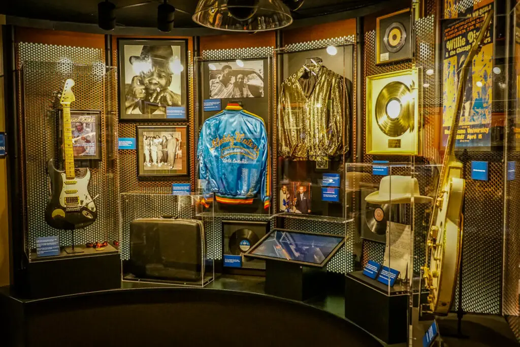 Interior view of a music museum display, showcasing a variety of memorabilia including a black and white electric guitar, several framed photographs of musicians, a shiny gold jacket, a blue satin jacket with 'Muddy Waters' text, and multiple framed gold records.