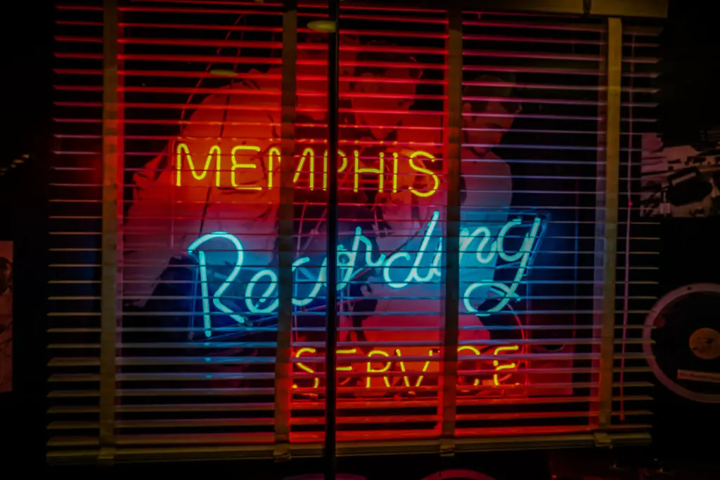 Neon sign through window blinds reading "Memphis Recording Service" in red and blue, with a faint silhouette of a person in the background. Sun Studio is one of the best museums in Memphis, TN.
