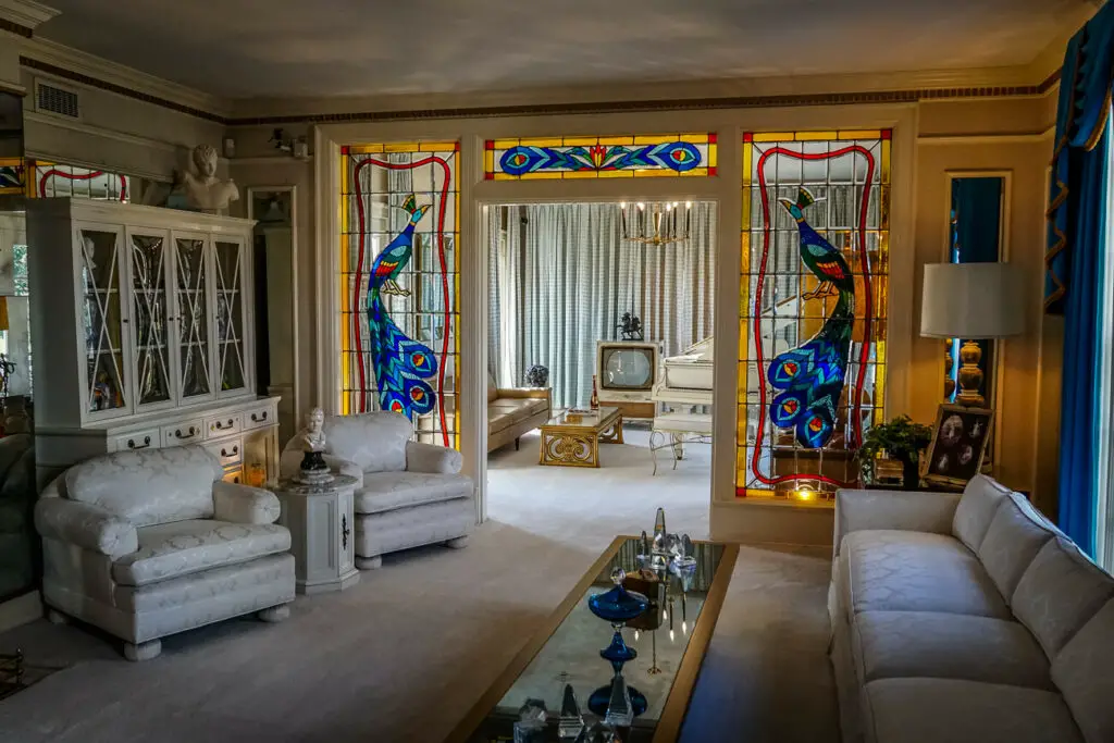 Opulent living room featuring white sofas, a mirrored coffee table, and extravagant stained glass peacock designs on either side of a doorway. Elvis Presley's former home, Graceland, is one of the best museums in Memphis - take a sneak peek into his once-opulent lifestyle.