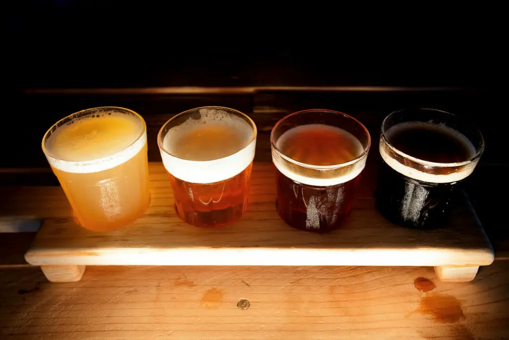 Four glasses of beer in a tasting flight ranging from light to dark, served on a wooden paddle, representing Fredericksburg's brewery offerings.