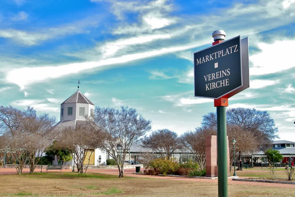 Directional sign pointing to 'Marktplatz Vereins Kirche' with a white historic church in the background against a blue sky with streaky clouds. This building can be found in Fredericksburg TX.