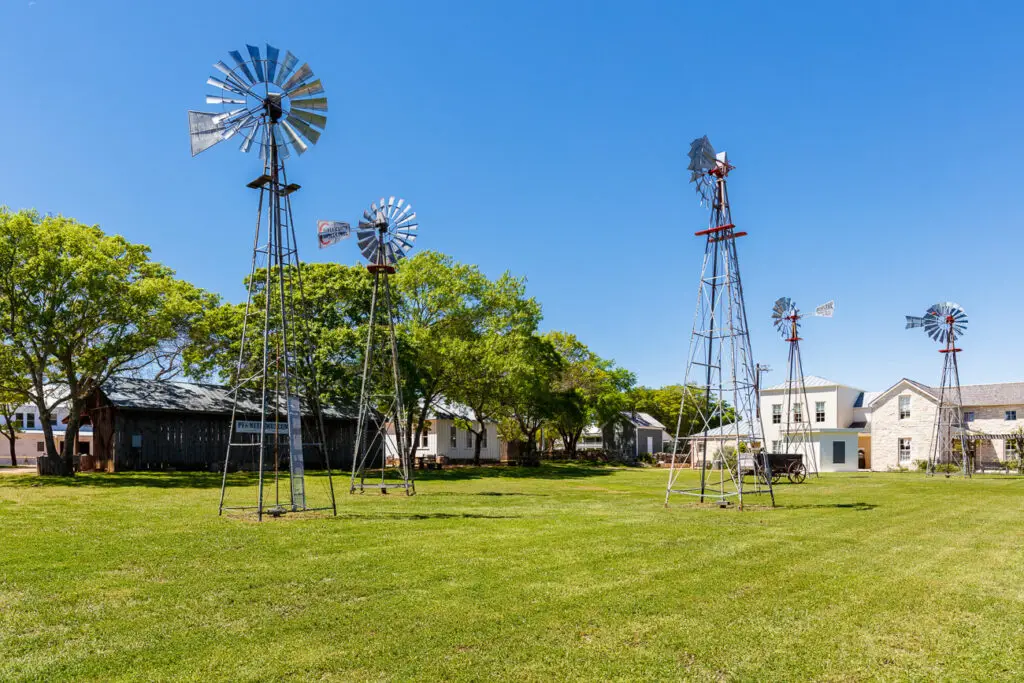 Three metal windmills standing in an open grassy field with historic buildings in the distance under a clear blue sky. The Pioneer Museum is an interesting places to visit in Fredericksburg Texas.