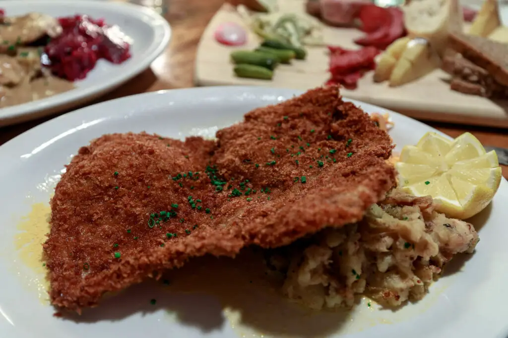 Close-up of a traditional Wiener Schnitzel served with lemon and potato salad on a plate, with other German dishes in the background. There are several German restaurants in Fredericksburg TX to try!