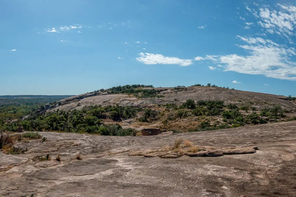 Expansive view of Enchanted Rock with a bare, dome-shaped granite hill rising under a blue sky with fluffy clouds. Hiking Enchanted Rock is one of the best things to do in Fredericksburg TX in a weekend.