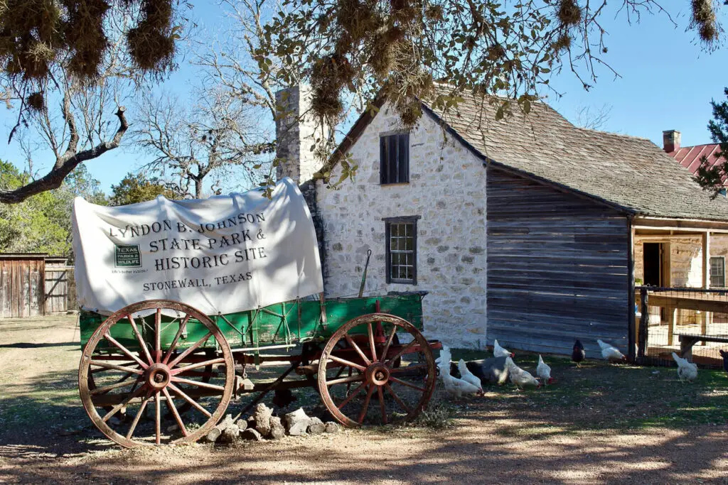 Old-fashioned covered wagon with 'Lyndon B. Johnson State Park & Historic Site, Stonewall, Texas' sign, next to a stone building and free-roaming chickens.